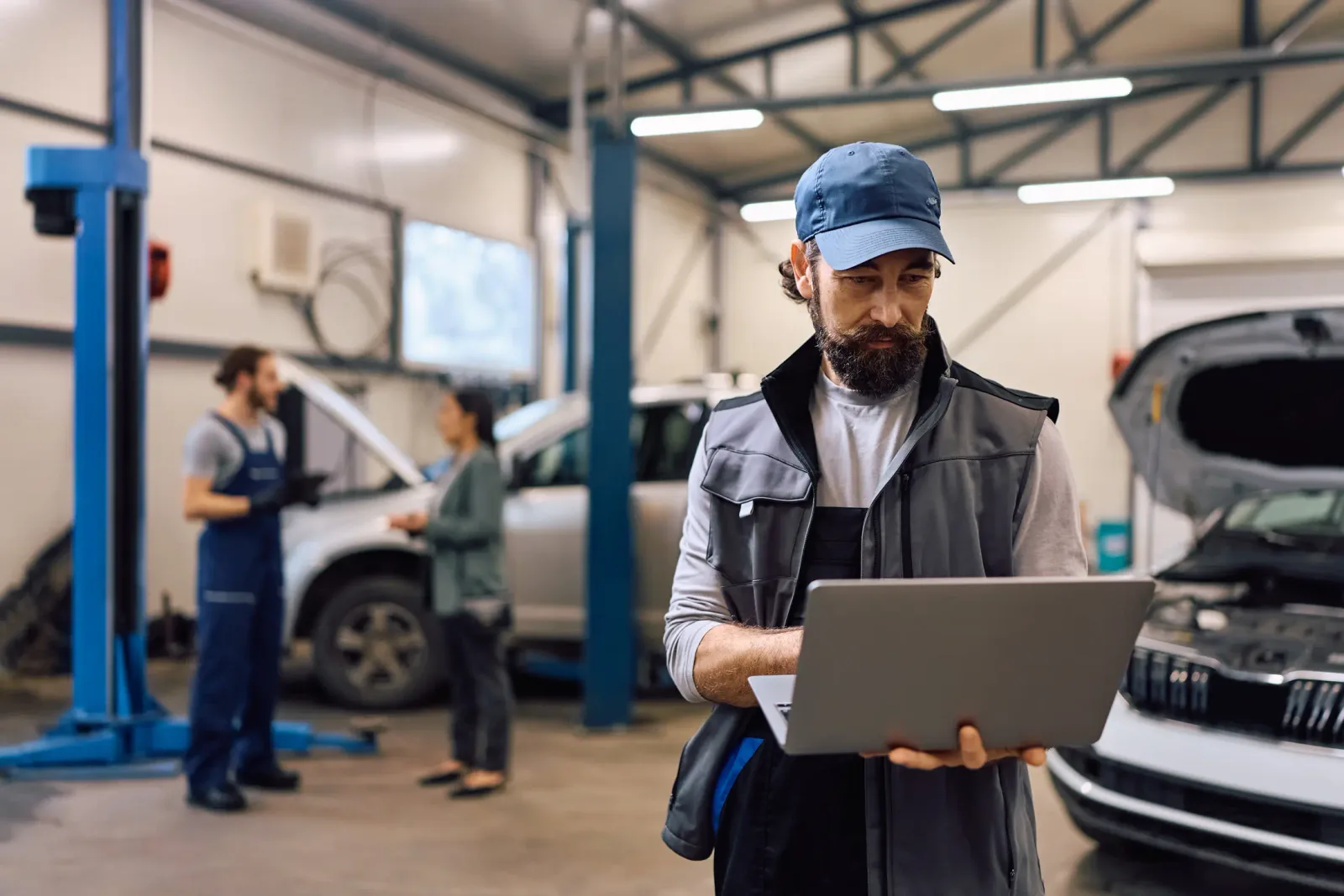 An auto shop: a mechanic with a laptop in the foreground while a colleague talks to a customer at a car on the lift